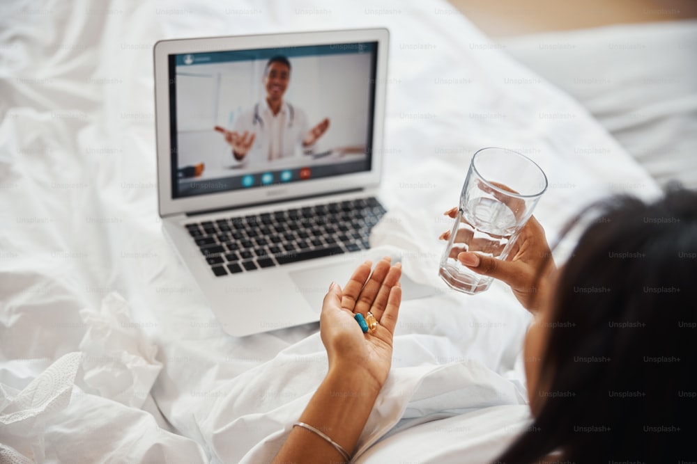 Gros plan d’une femme tenant des médicaments et un verre d’eau alors qu’elle est allongée dans son lit avec un ordinateur portable et parle avec un médecin en ligne