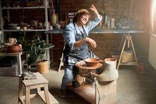 Nice female potter in apron covering face from sun and smiling while making clay bowl on pottery wheel in studio
