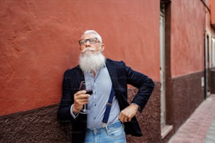 Handsome fashionable senior man with white beard and mustache holding glass of red wine and thinking about something. Outdoor photo. Elegant pensioner posing.