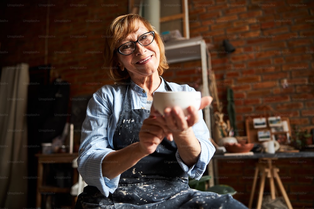 Artista de cerámica de la mujer mayor alegre que mira a la cámara y sonríe mientras sostiene un cuenco de barro hecho a mano