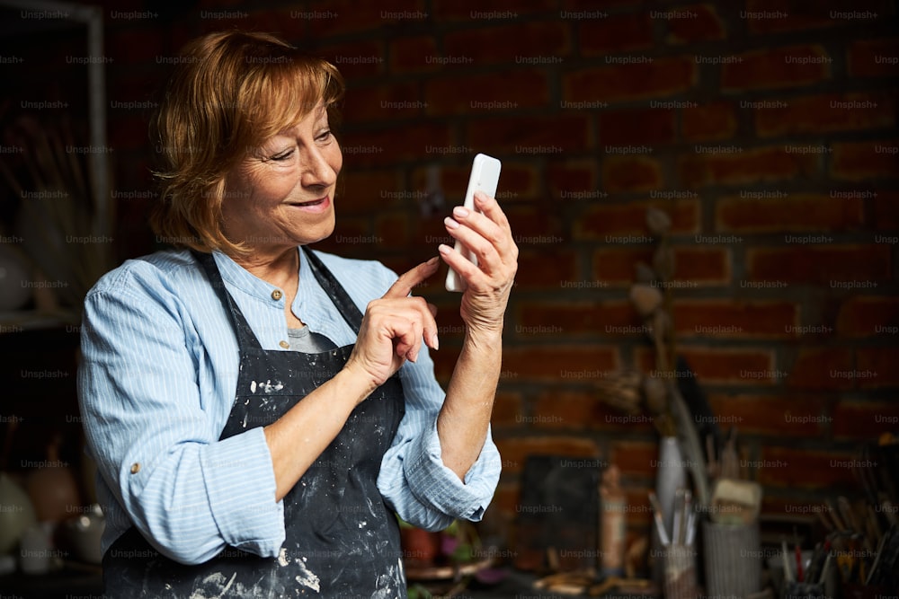 エプロンで素敵な女性の陶芸家がスマートフォンでメッセージをテキストメッセージで送信し、陶芸スタジオに立って微笑みます