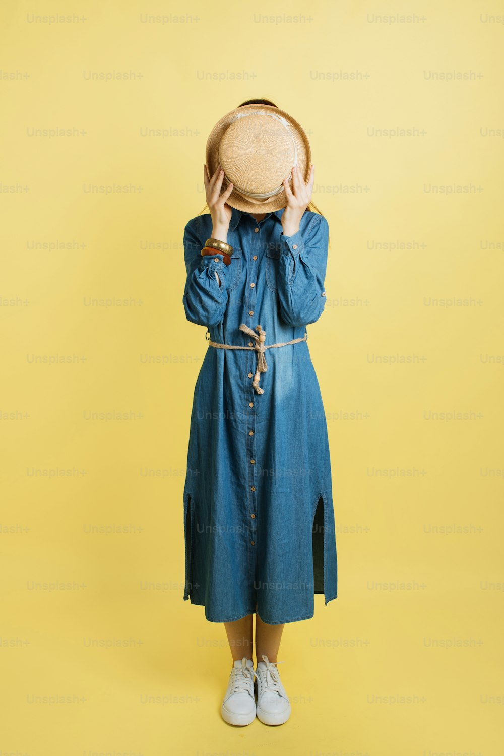 Portrait of attractive young trendy travel woman, wearing jean dress, hiding face with stylish summer straw hat, posing on camera in studio isolated over yellow background