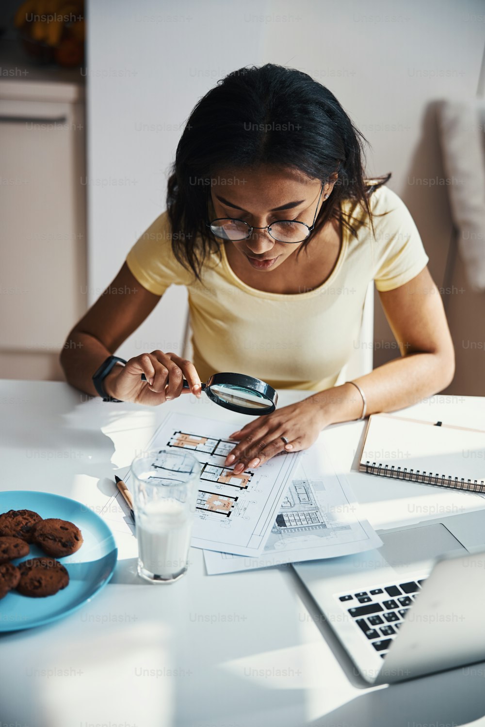 Charming young woman holding magnifying glass and analyzing building plan while sitting at the table and working on project