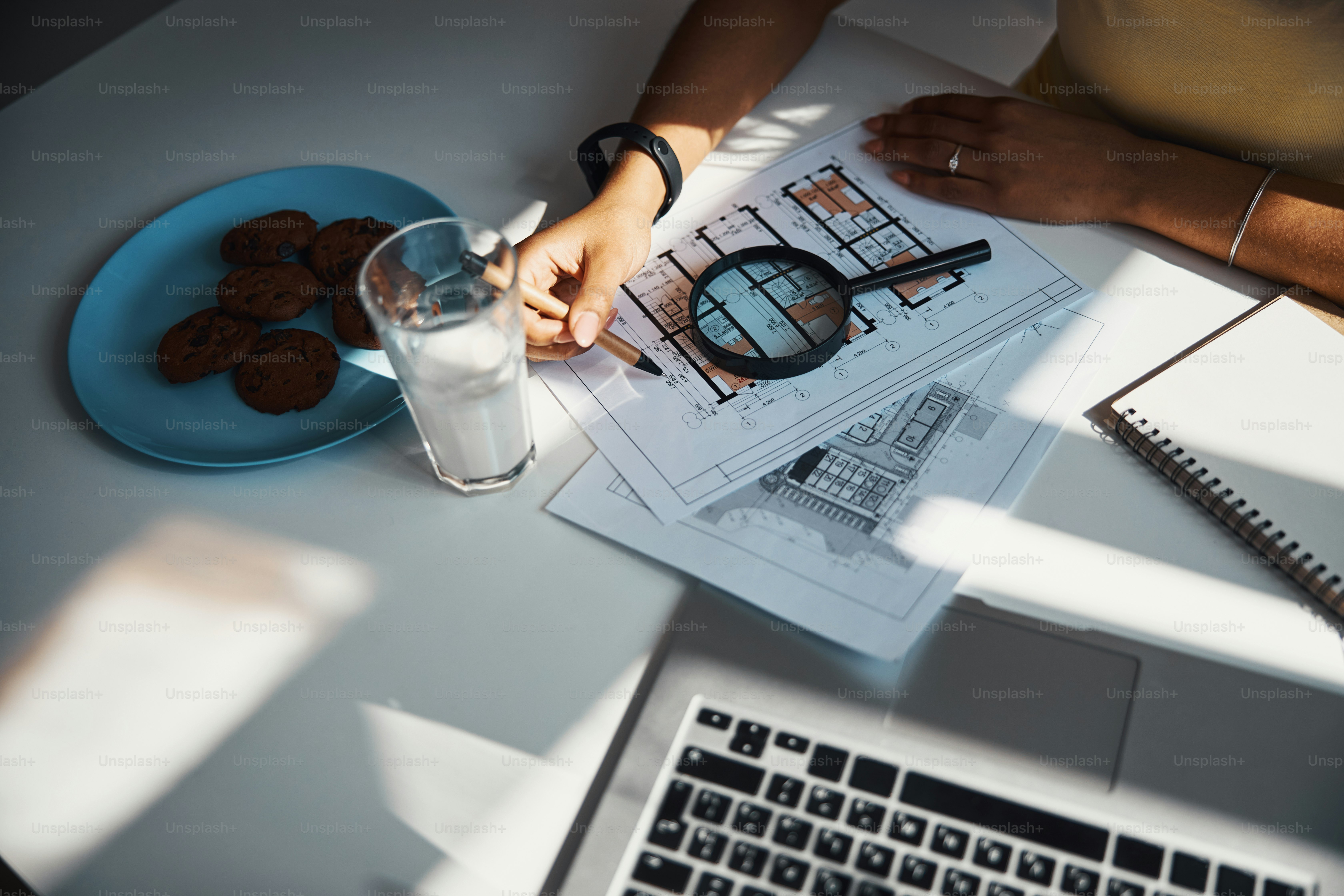 Close up of young woman with pen in hand working on architectural plan while sitting at the table with laptop, glass of milk and cookies