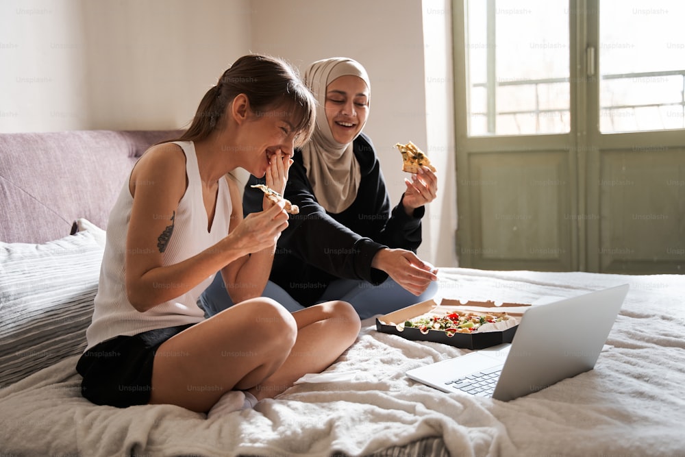 Spaß. Volle Ansicht der beiden Mädchen, die frische Pizza essen und laut lachen, während sie lustige Filme im Schlafzimmer zu Hause sehen. Archivfoto