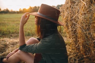 Beautiful stylish woman in hat enjoying sunset, sitting at haystacks in evening summer field. Atmospheric tranquil moment in countryside. Young fashionable female relaxing at hay bale in sunshine