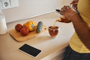 スマートフォンと野菜を持ってキッチンカウンターのそばに立っている間、ティーポットを持ち、マグカップに温かい飲み物を注ぐ若い女性の接写