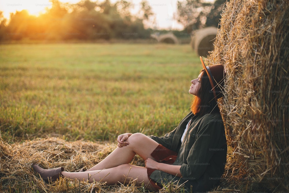 Hermosa mujer elegante con sombrero que disfruta de la puesta de sol, sentada en pajares en el campo de verano de la tarde. Momento de tranquilidad atmosférica en el campo. Joven mujer de moda relajándose en la paca de heno bajo el sol