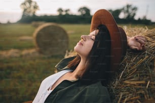 Beautiful stylish woman with herb in mouth and in hat relaxing on haystack in summer field. Portrait of attractive young female enjoying evening at hay bale. Atmospheric tranquil moment
