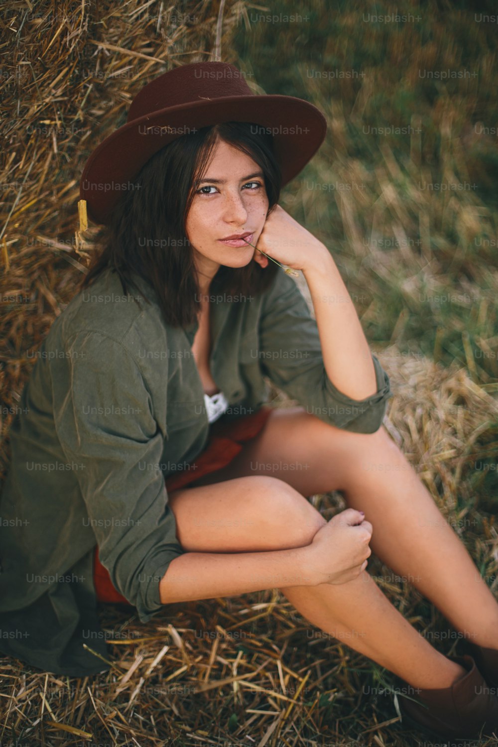 Beautiful stylish woman with herb in mouth and in hat sitting at haystack in summer evening field. Portrait of sexy young female relaxing at hay bale, summer vacation in countryside.