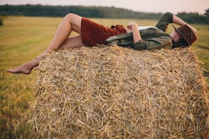 Beautiful carefree woman in hat lying on haystack enjoying evening in summer field. Young happy female relaxing on hay bale in countryside. Atmospheric tranquil moment
