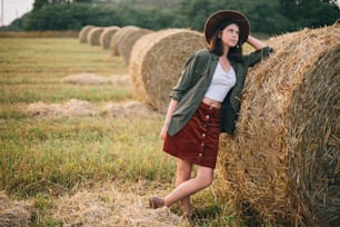 Stylish woman in hat standing at hay bale in summer evening in field. Atmospheric tranquil moment. Young fashionable female relaxing at haystack, summer vacation in countryside. Rural slow life
