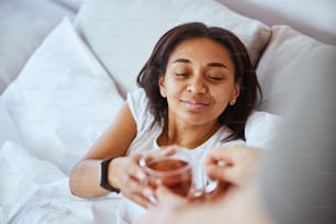 Hermosa mujer afroamericana acostada en la cama y sonriendo mientras el novio le da una deliciosa bebida caliente