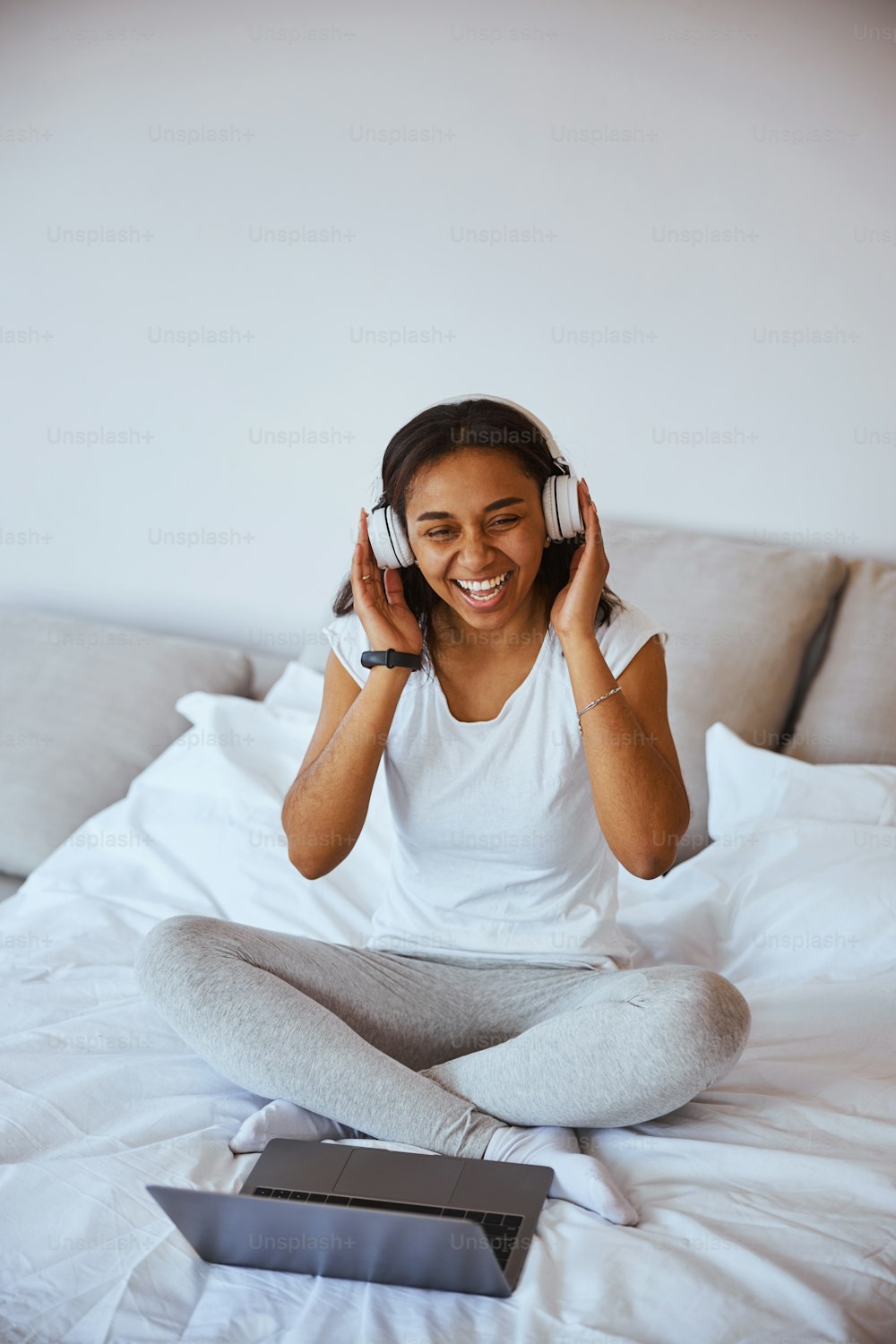 헤드폰을 끼고 좋아하는 노래를 즐기고 노트북으로 침대에 앉아 웃고 있는 아름다운 아프리카계 미국인 여성