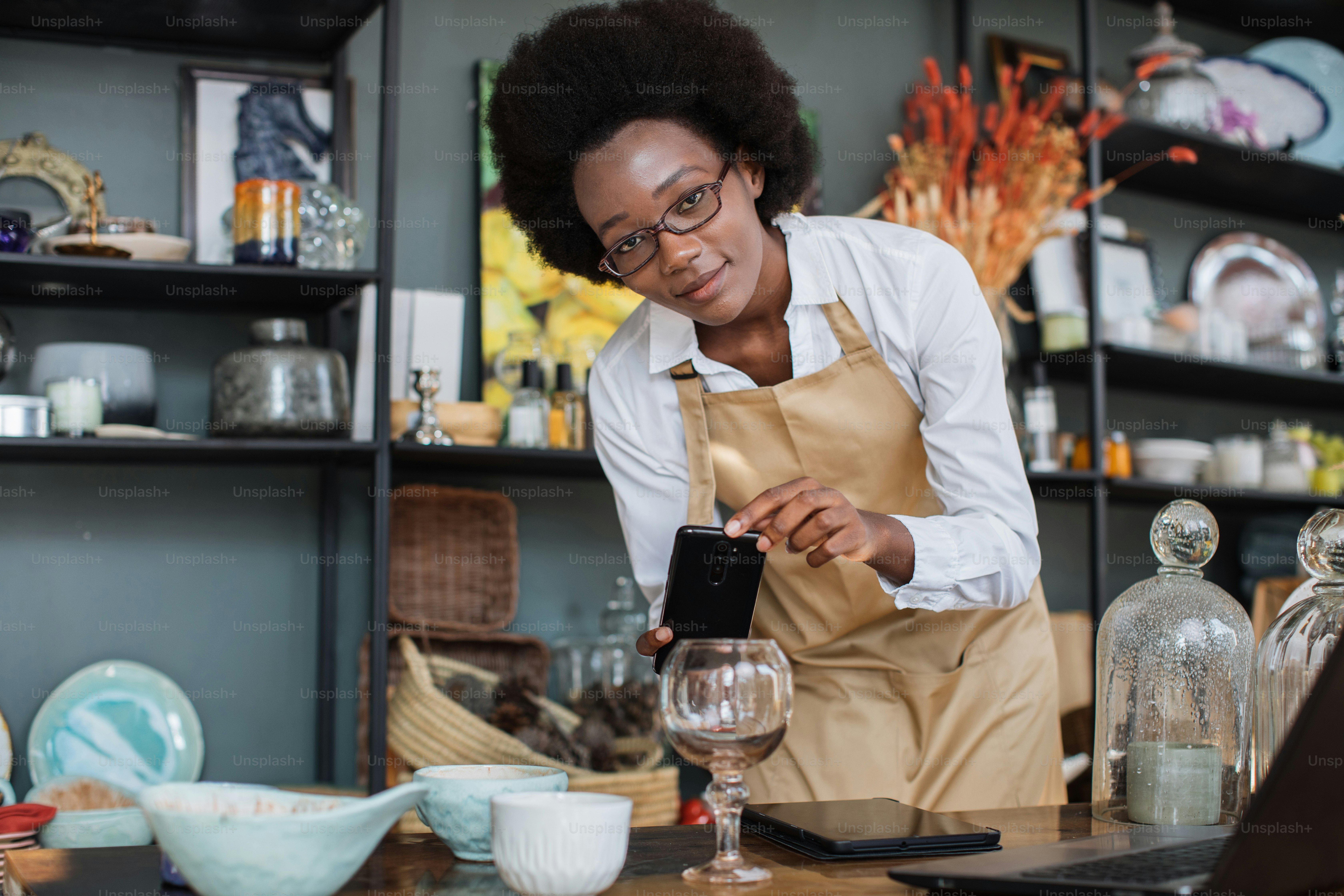 Nahaufnahme einer afrikanischen Verkäuferin mit Brille, die Dekorationen und Glas im Geschäft auf einem modernen Smartphone fotografiert und einen Online-Katalog mit einem drahtlosen Laptop erstellt.