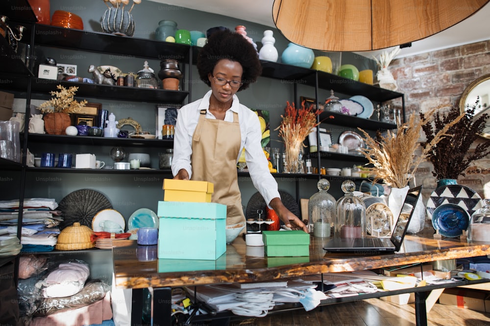 Vendedora afroamericana calificada en anteojos empacando cajas con varios recuerdos para los clientes. Hermosa mujer que trabaja en una tienda moderna con decoración.