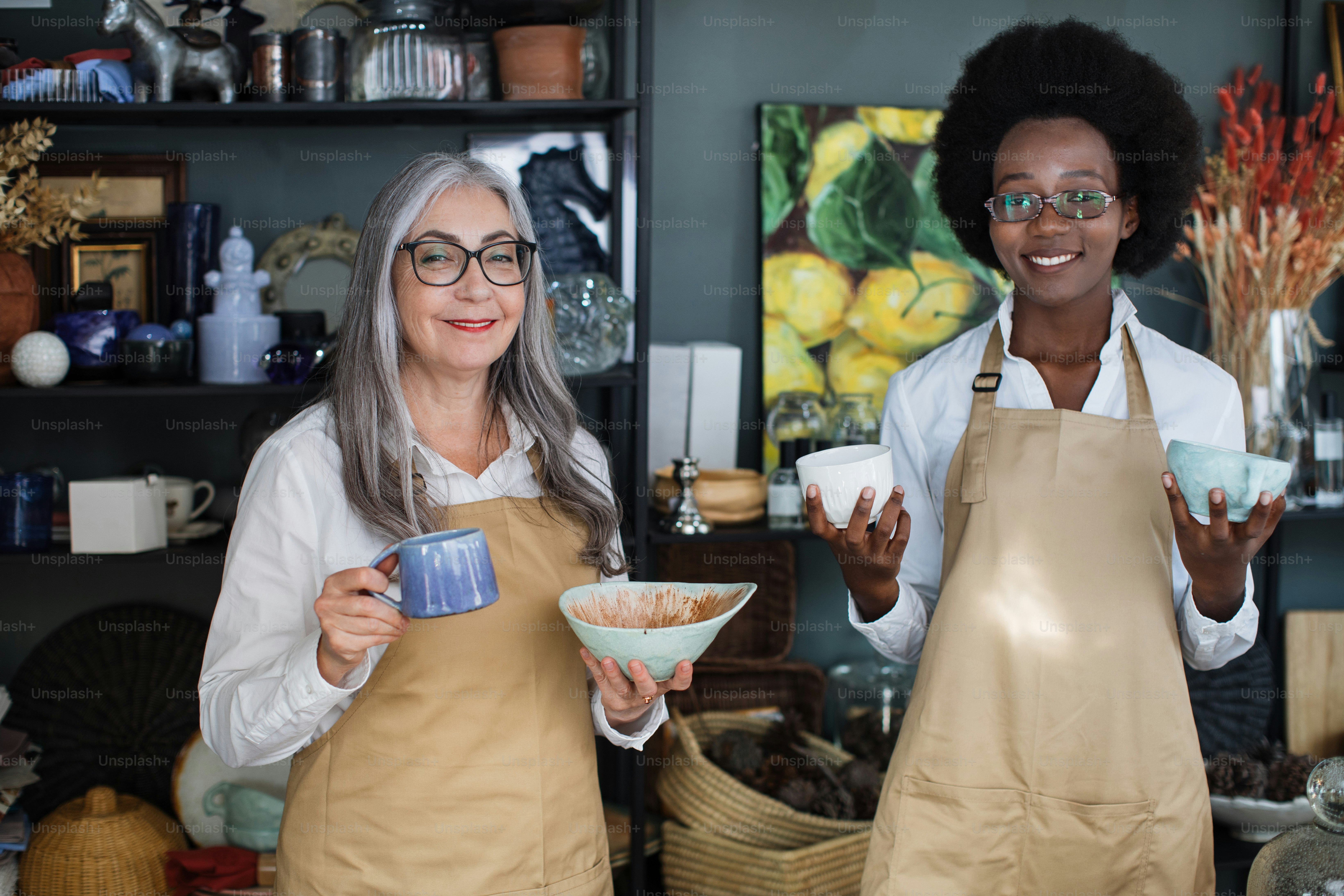 Two diverse saleswomen in beige apron and eyeglasses posing together at store an holding various cups in hands. Happy people working at decor shop.