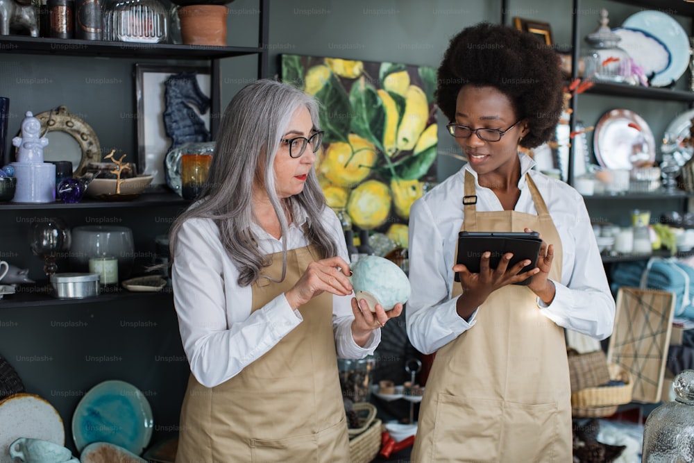 Young african woman and mature caucasian woman standing at decor shop and using digital tablet for receiving goods. Working process at store.