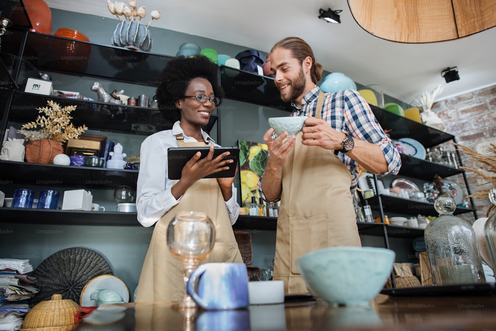 Two multiracial partners using digital tablet while receiving goods at decor store. African woman and caucasian man wearing beige aprons, working together.