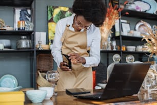 African saleswoman in eyeglasses taking picture of all assortment of decor shop on modern smartphone. Pretty woman in apron creating online catalogue using wireless laptop.