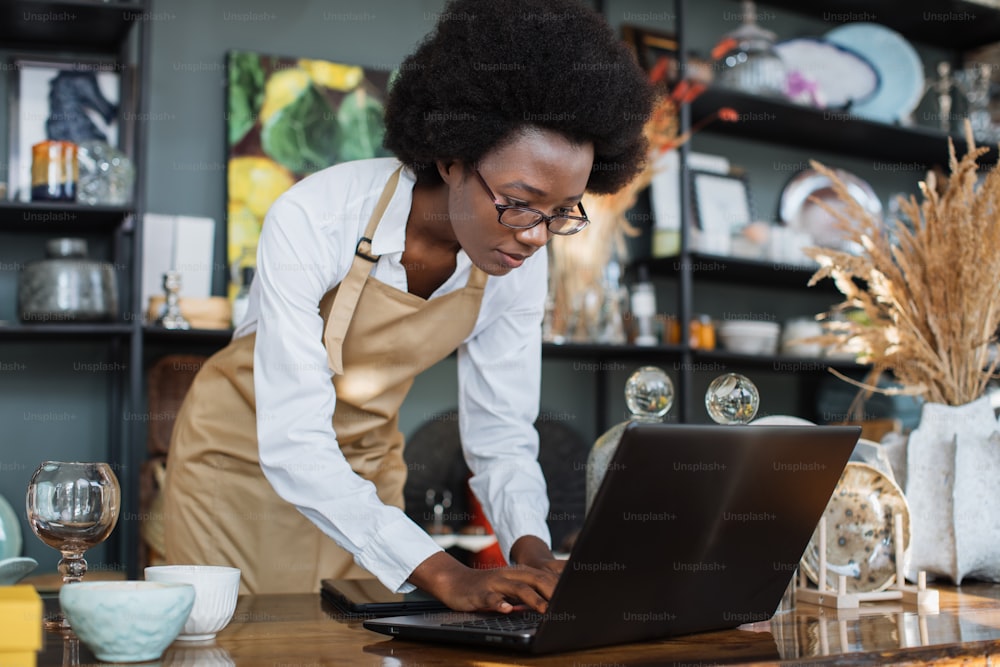Focused afro american woman in eyewear and apron using wireless laptop during working process at shop with various decor. Creating website for online shopping.