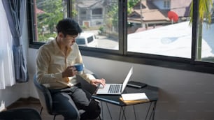 Casual businessman drinking coffee and surfing internet on laptop computer at office in the morning.