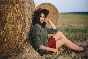 Bella donna elegante con l'erba in bocca ed in cappello seduta al pagliaio nel campo di sera d'estate. Giovane femmina sexy che si rilassa alla balla di fieno, vacanza estiva in campagna.