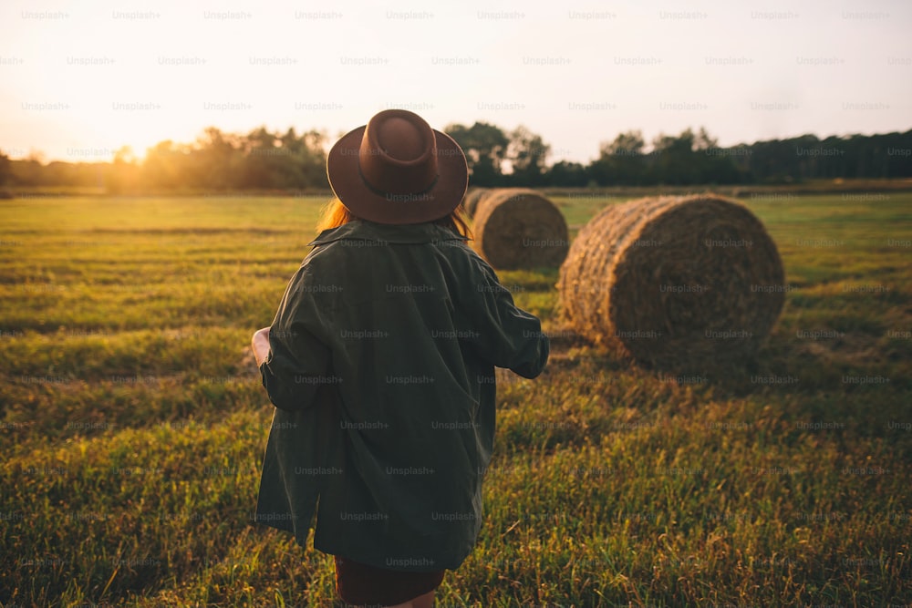 夏の野原の夕暮れの光の中で干し草の山を歩く帽子をかぶった美しいスタイリッシュな女性。田舎の雰囲気の静かなひととき。暖かい日差しの中で干し草の俵で夜を楽しむ若い女性