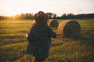 Belle femme élégante en chapeau marchant à des meules de foin dans la lumière du coucher du soleil dans le champ d’été. Moment tranquille atmosphérique à la campagne. Jeune femelle profitant d’une soirée à une balle de foin sous un soleil chaud