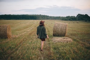 Beautiful stylish woman in hat walking at hay bales in summer evening field. Happy young female relaxing at haystacks, vacation in countryside. Tranquility, countryside slow life