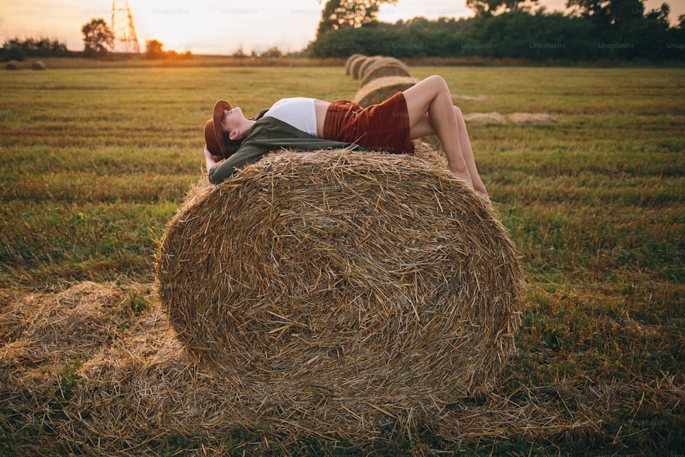 Beautiful carefree woman in hat lying on haystack in sunset light enjoying evening in summer field. Young happy female relaxing on hay bale in countryside. Atmospheric tranquil moment