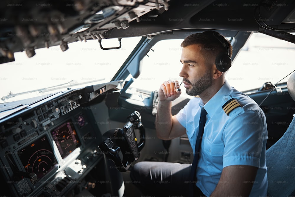 Jovem piloto caucasiano franzindo a testa enquanto está sentado em um cockpit durante o dia e se comunicando através do transmissor