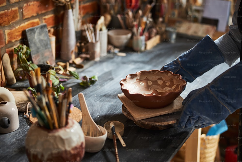 Close up of ceramic artist in gloves holding earthenware bowl while working in art pottery studio
