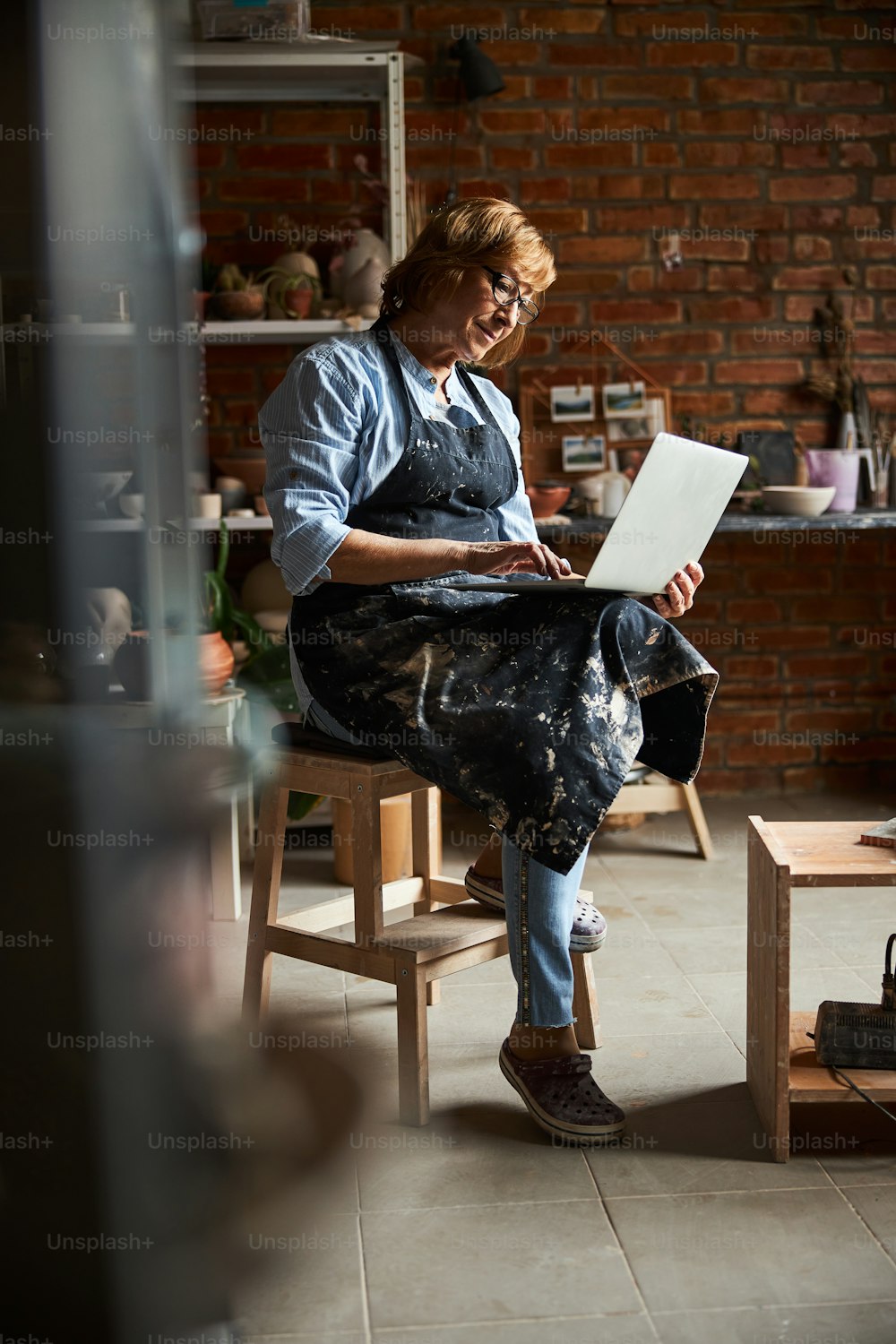 エプロンで魅力的な女性の陶芸家がラップトップのキーボードでタイピングし、陶器スタジオのスツールに座って微笑む