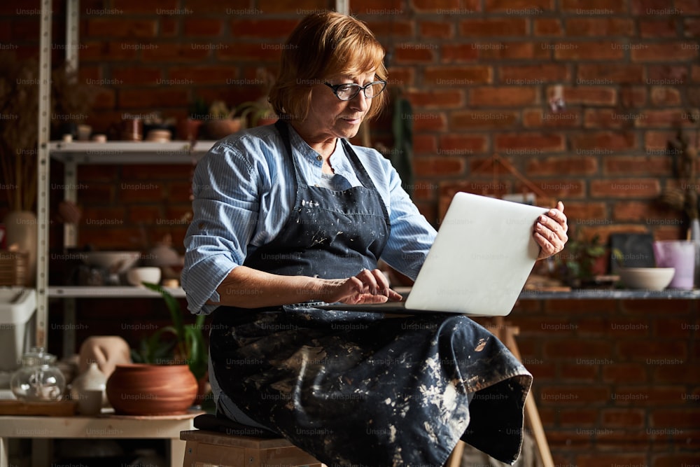 Charming female potter in apron sitting on stool and typing on notebook keyboard while working in pottery studio