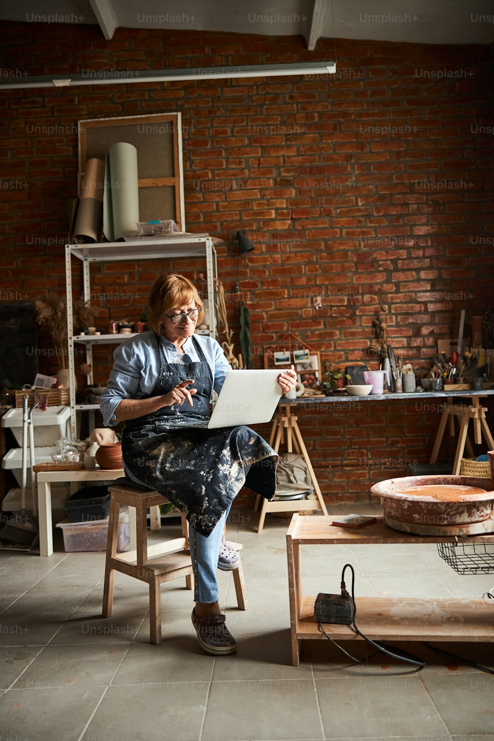 Encantadora anciana artista de cerámica viendo video en computadora portátil y sonriendo mientras está sentada en un taburete en un estudio de cerámica