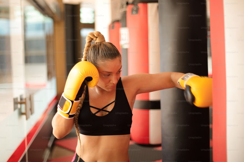 Strong young lady with boxing gloves, punching bag in gym alone.