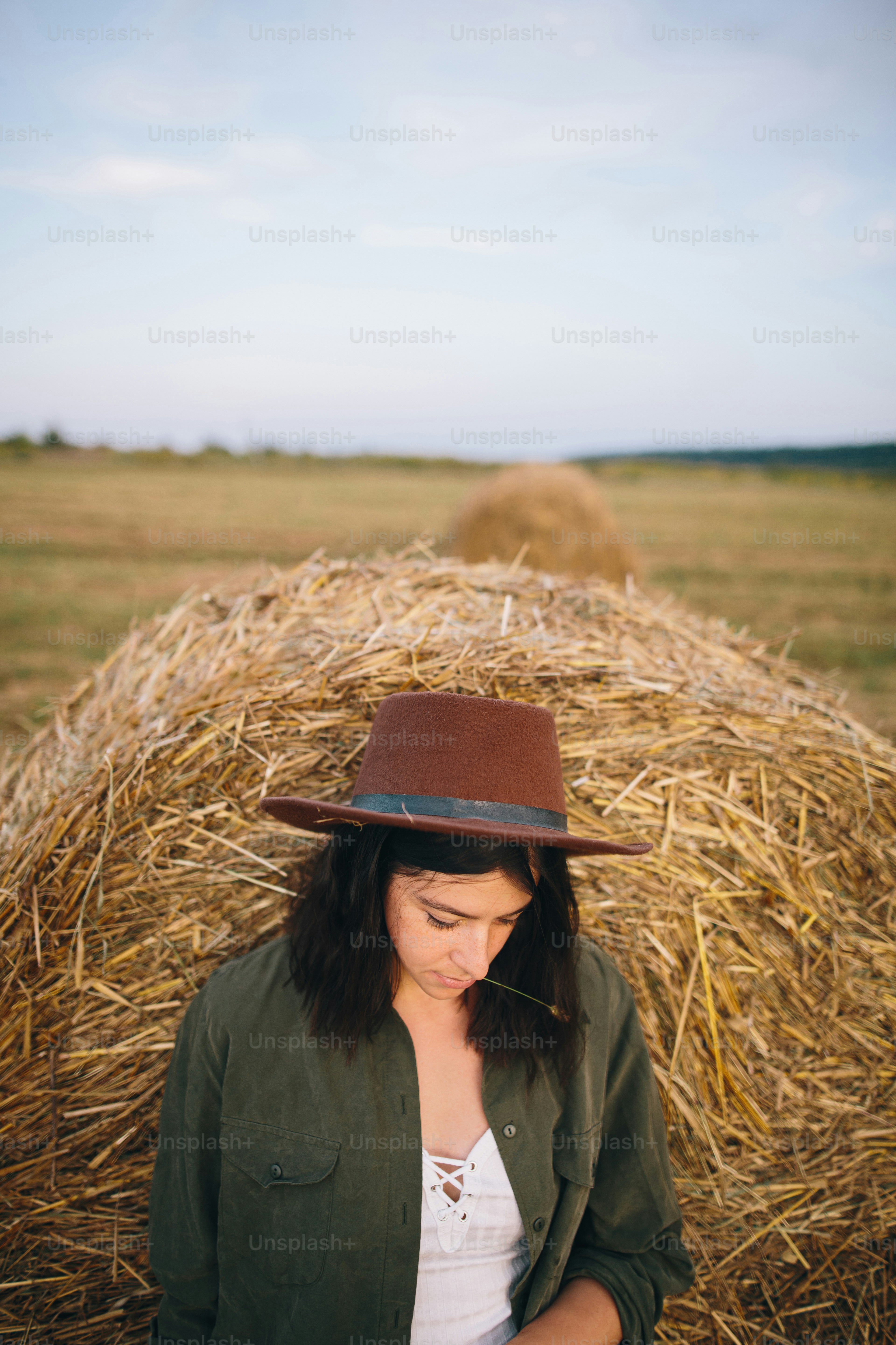 Stylish woman in hat standing at hay bale in summer evening in field. Atmospheric tranquil moment. Young fashionable female relaxing at haystack, summer vacation in countryside. Rural slow life