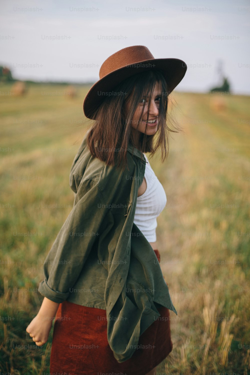 Beautiful carefree woman in hat walking and smiling in evening summer field. Young happy stylish female relaxing in countryside, enjoying evening. Atmospheric moment.