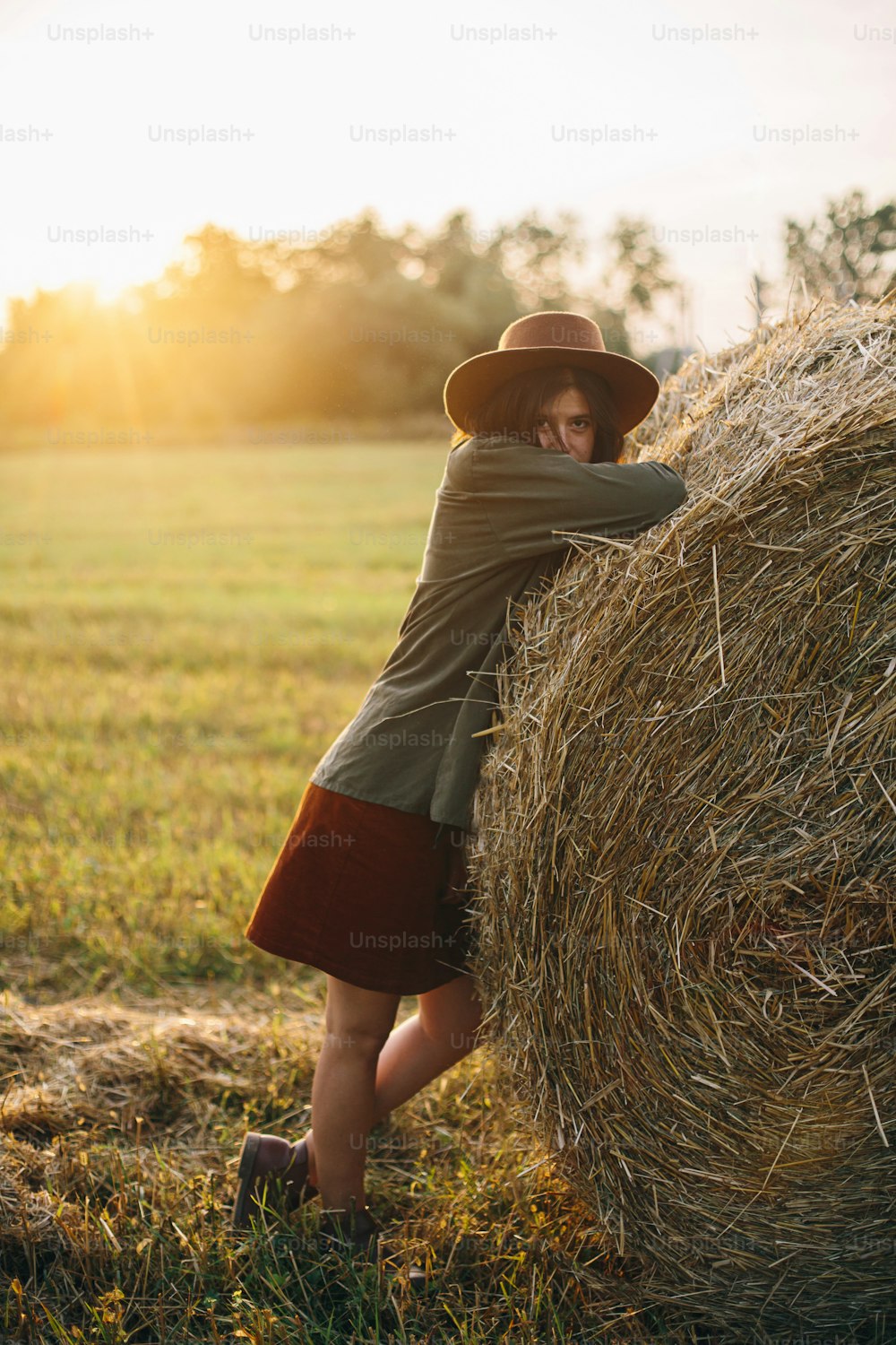 Beautiful stylish woman in hat posing at haystacks in sunset light in summer field. Atmospheric tranquil moment in countryside. Young female relaxing at hay bale in evening warm sunshine