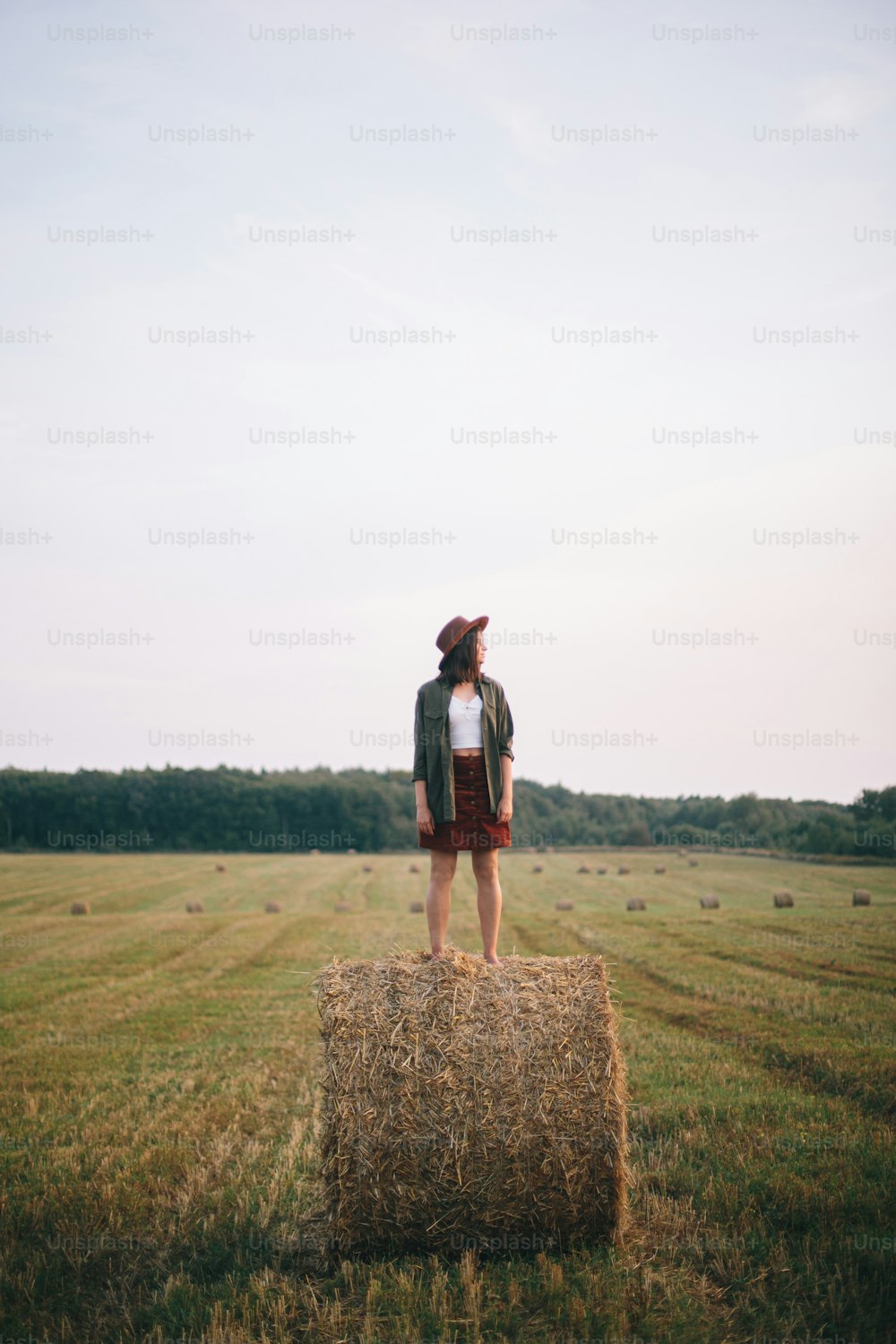 Beautiful carefree woman in hat standing on haystack enjoying evening in summer field. Young happy female relaxing on hay bale in countryside. Atmospheric tranquil moment