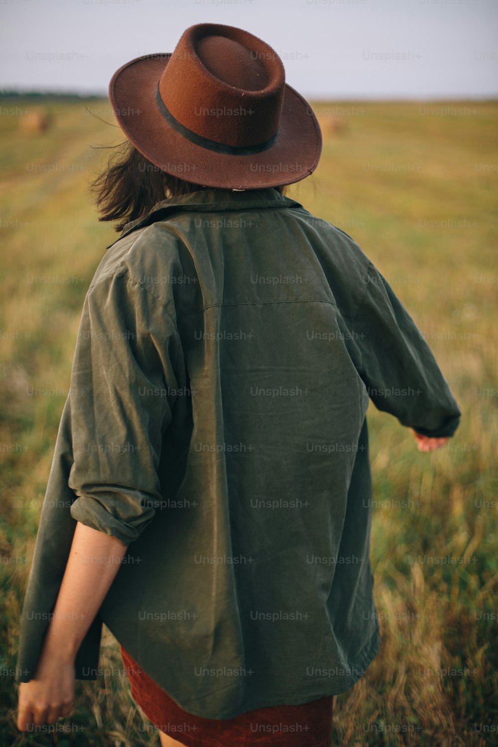 Beautiful carefree woman in hat walking in evening summer field. Young happy stylish female relaxing in countryside, dancing and enjoying evening. Atmospheric moment. Back view