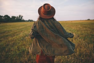 Beautiful carefree woman in hat walking in evening summer field. Young happy stylish female relaxing in countryside, dancing and enjoying evening. Atmospheric moment. Back view