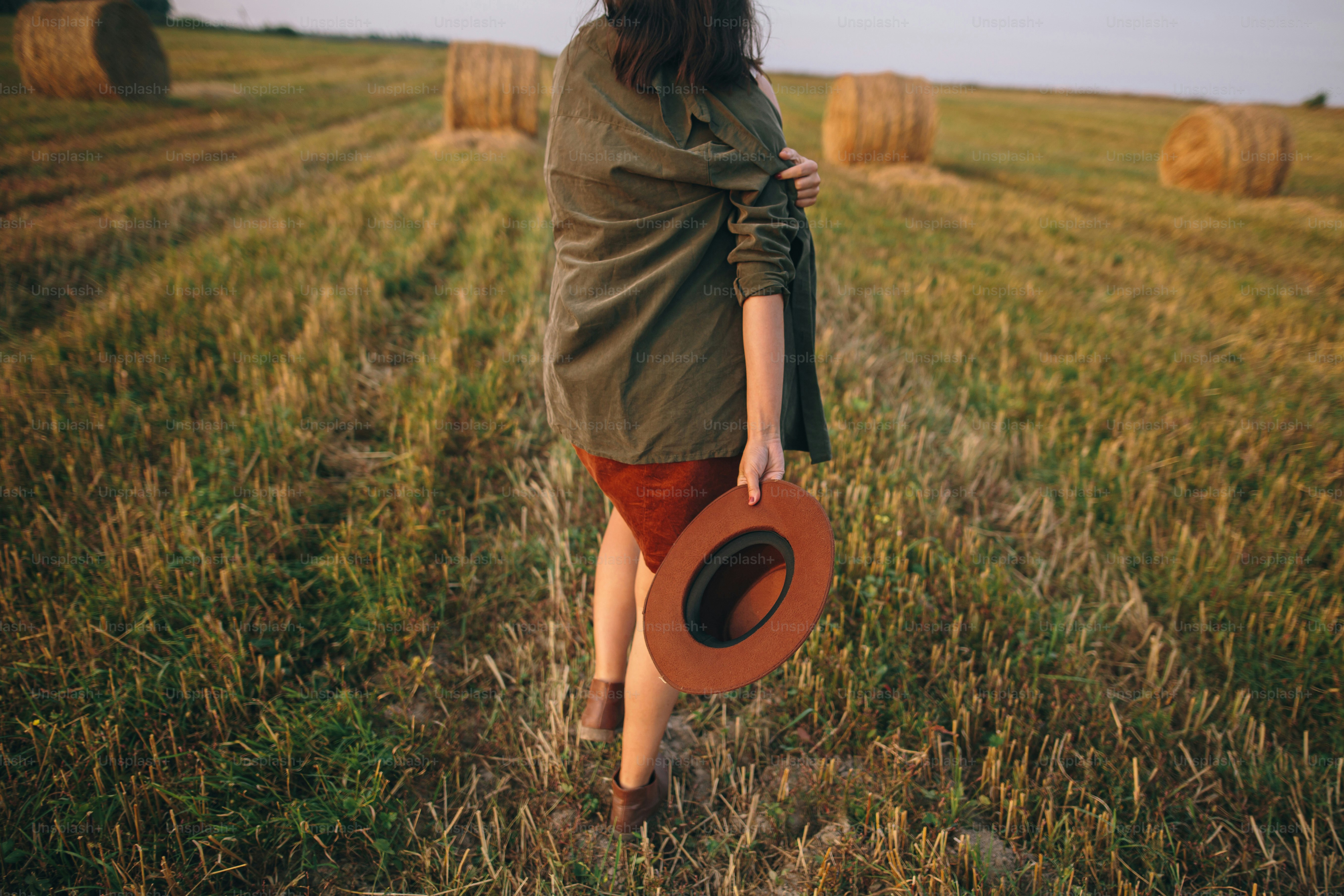 Beautiful carefree woman in hat walking in evening summer field. Young happy stylish female relaxing in countryside, dancing and enjoying evening. Atmospheric moment. Back view