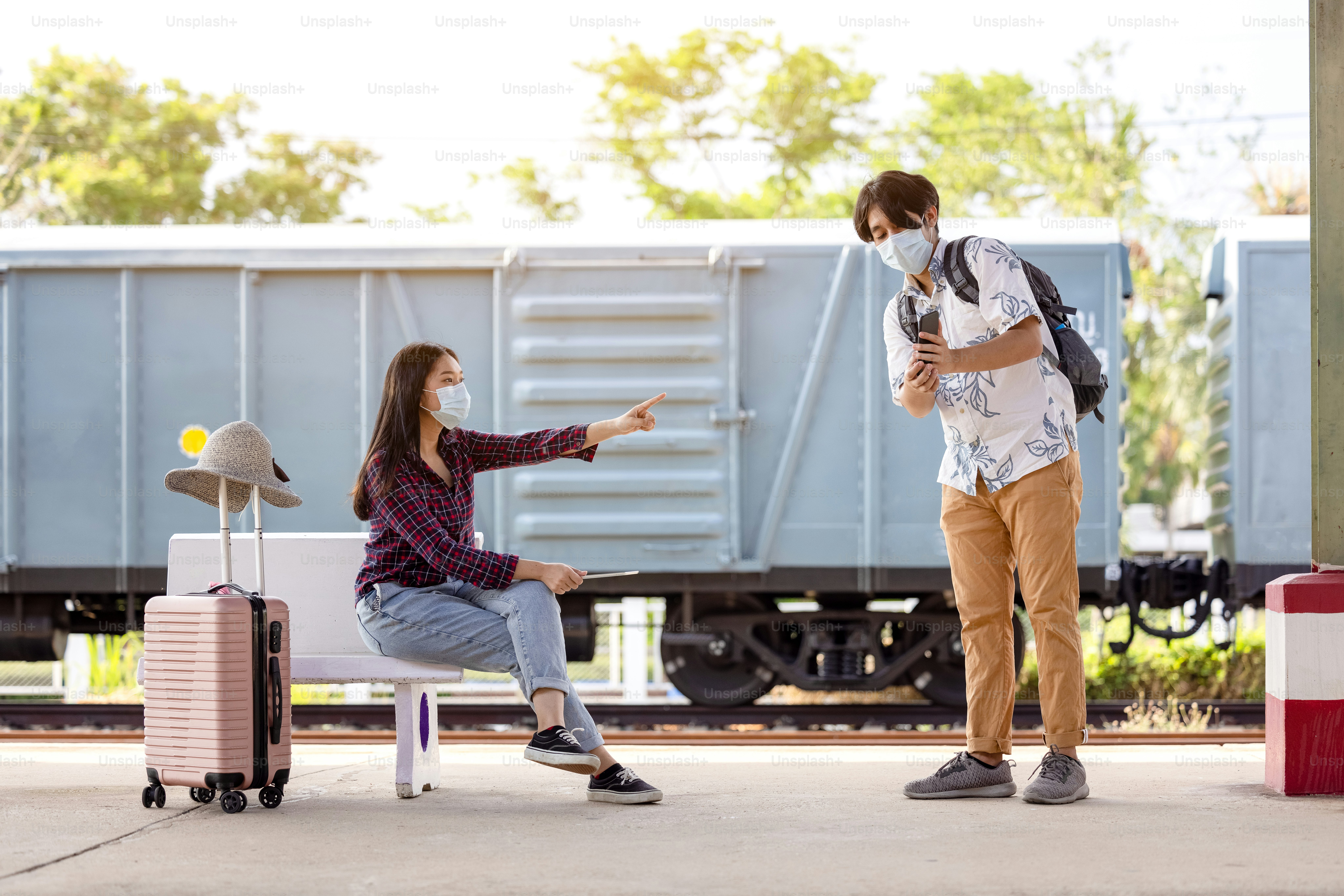 Young male traveler backpack with mask and mobile in hand asking way for help from woman sitting and pointing on stairs at subway, covid distance