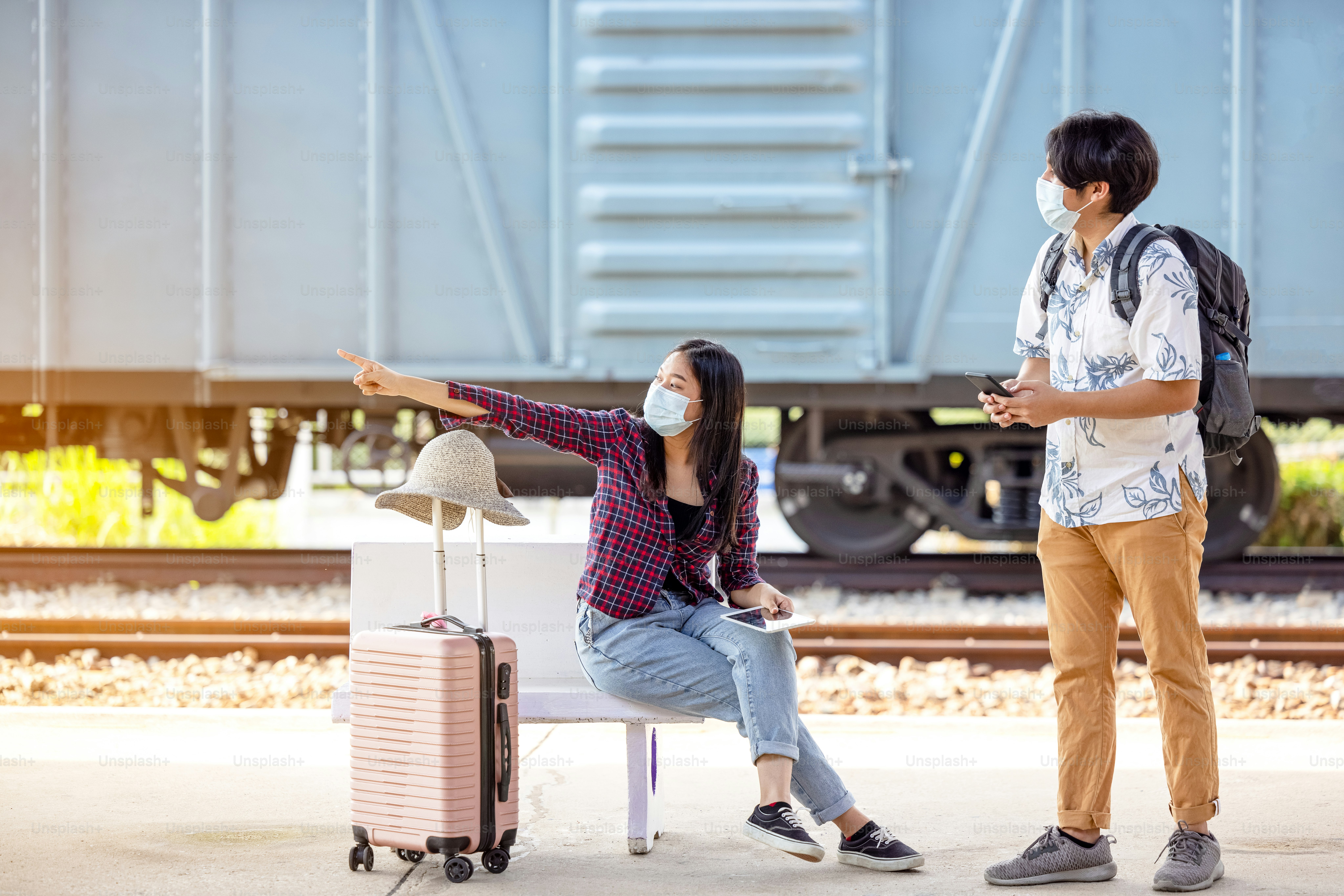 Young male traveler backpack with mask and mobile in hand asking way for help from woman sitting and pointing on stairs at subway, covid distance