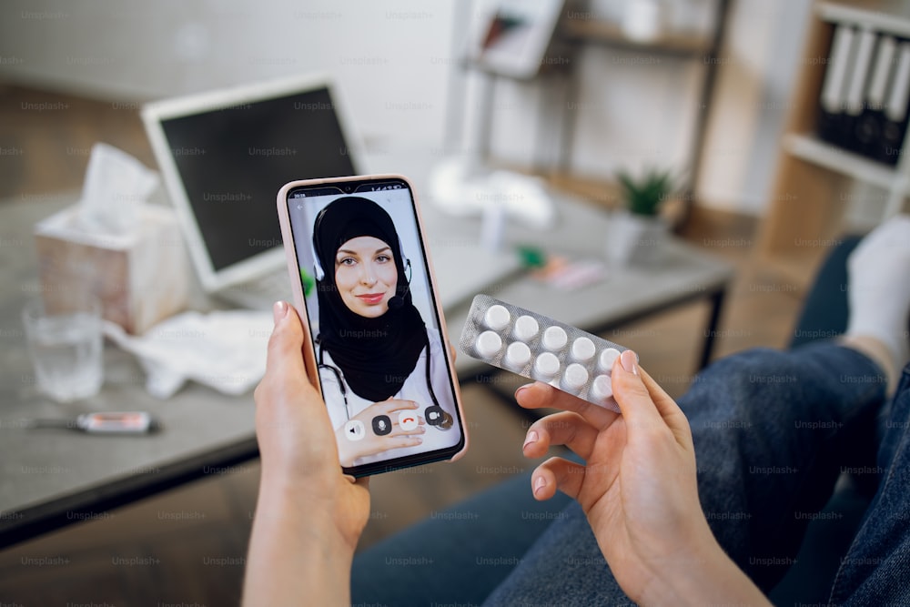 Telehealth care, treatment at home concept. Unrecognizable female patient communicating online with muslim medical worker, holding blister with white pills for treatment.