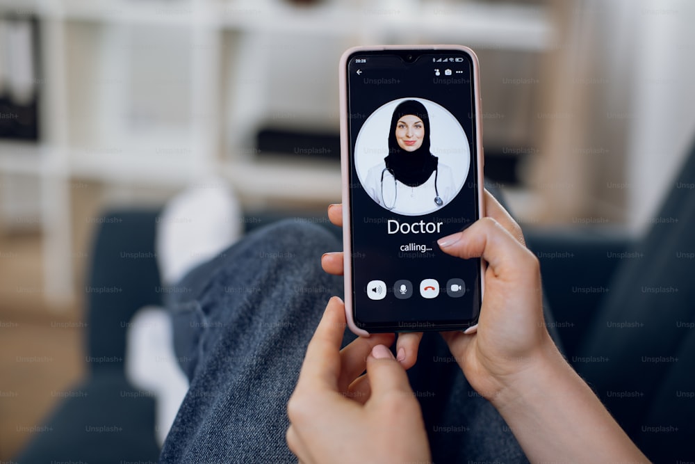 Close-up of hand of unrecognizable female patient, holding cell phone and having video conference with her Arabian muslim doctor in black hijab. Online consultation with doctor at home.