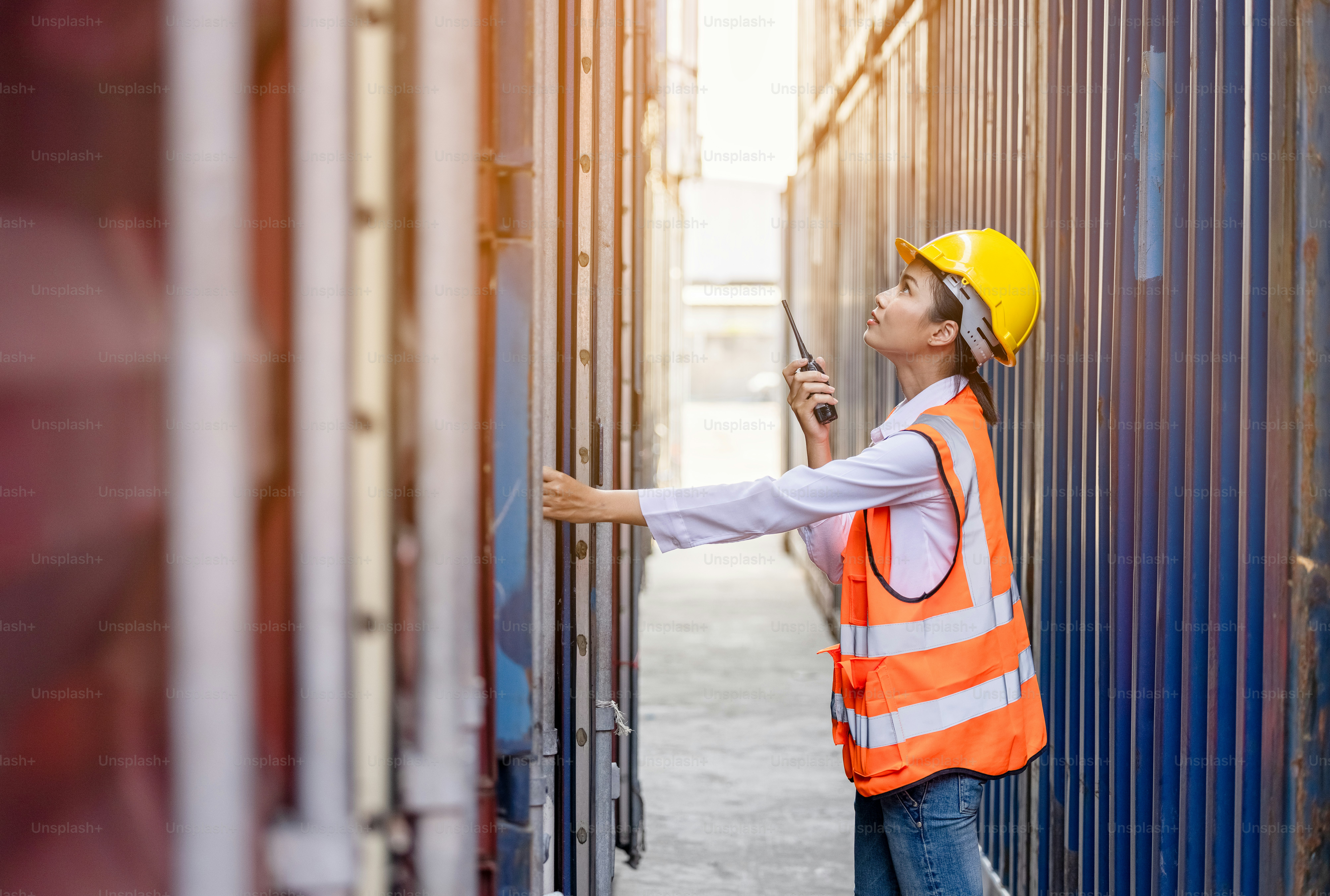 Porträt einer asiatischen Arbeiterin in Sicherheitsuniform, die mit Walkie-Talkie spricht, um die Arbeitsqualität im Containerlager zu kontrollieren. Arbeit für Logistik- und Versandgeschäfte.