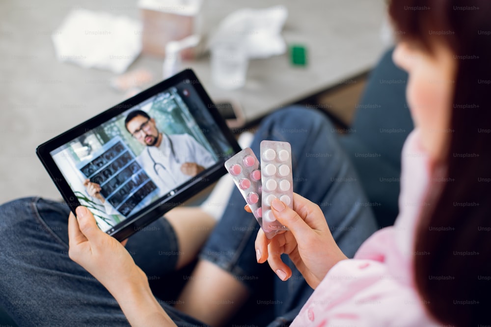 Concept de médecine, de technologie et de soins de santé. Jeune patiente, assise sur un canapé à la maison avec des pilules à la main, ayant une conversation vidéo avec un médecin masculin sur une tablette et consultant sur le traitement.